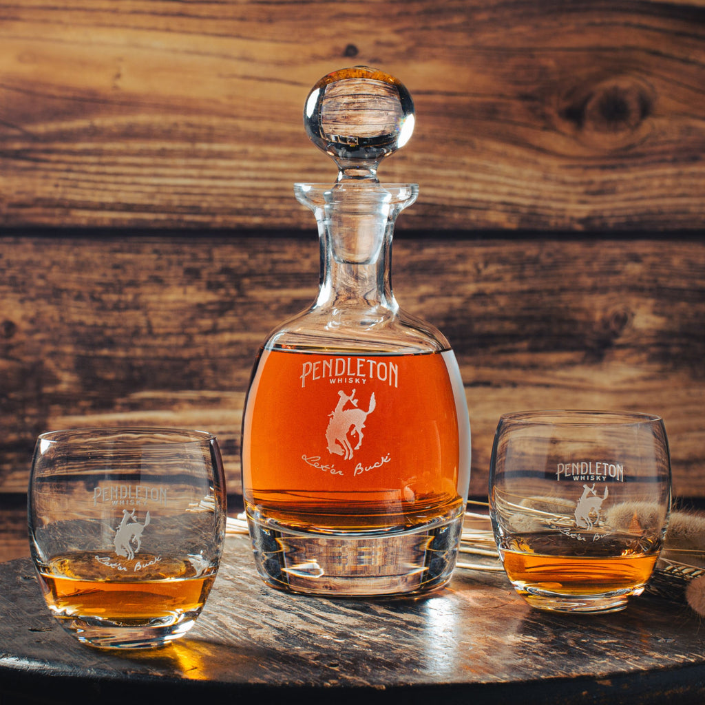 A crystal decanter with a Pendleton Whiskey logo and two rocks glasses, also with the Pendleton logo, on a wooden table.