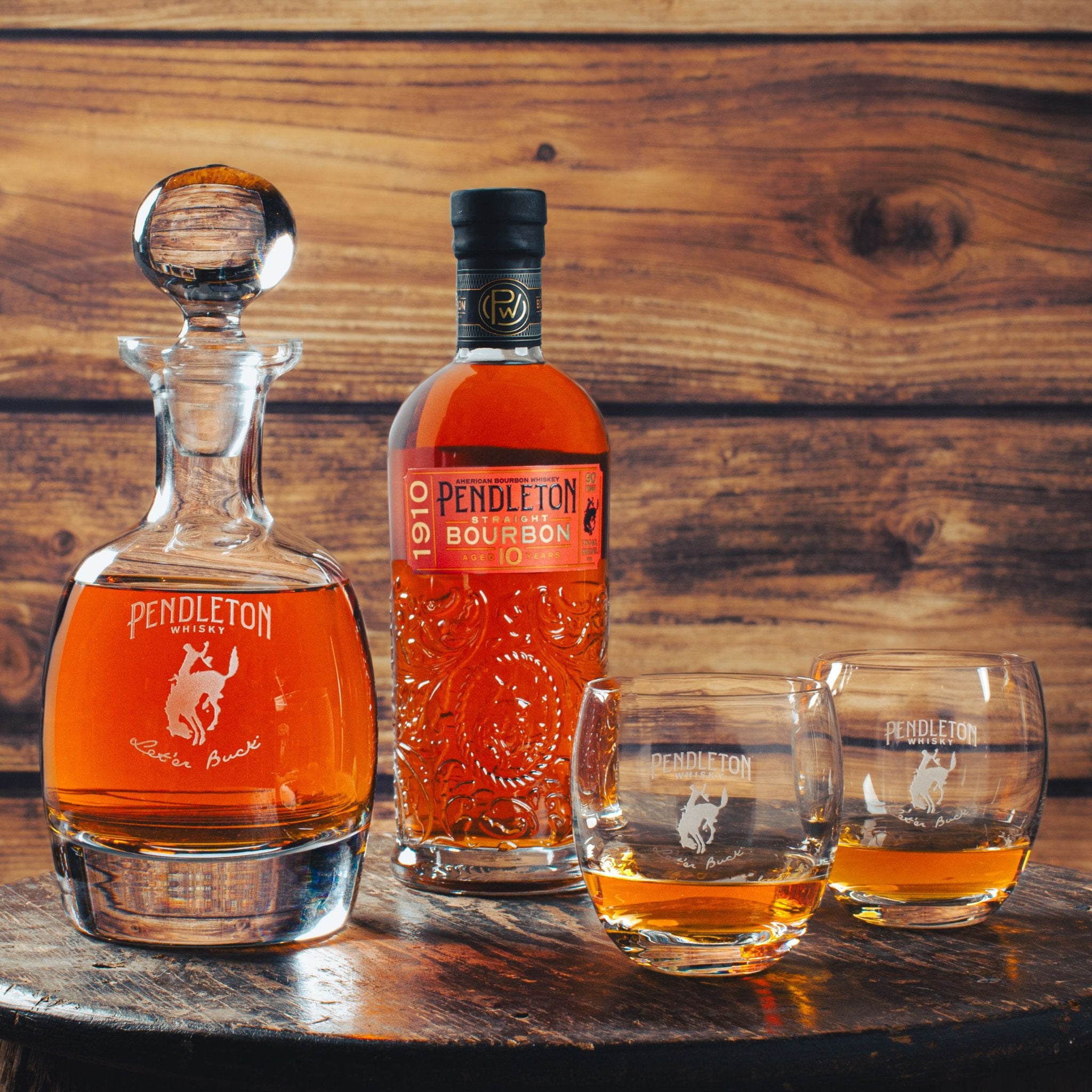 A bottle of Pendleton 1910 Straight Bourbon Whiskey, a decanter, and two rocks glasses on a wooden table. The bottle and glasses feature the Pendleton brand logo.